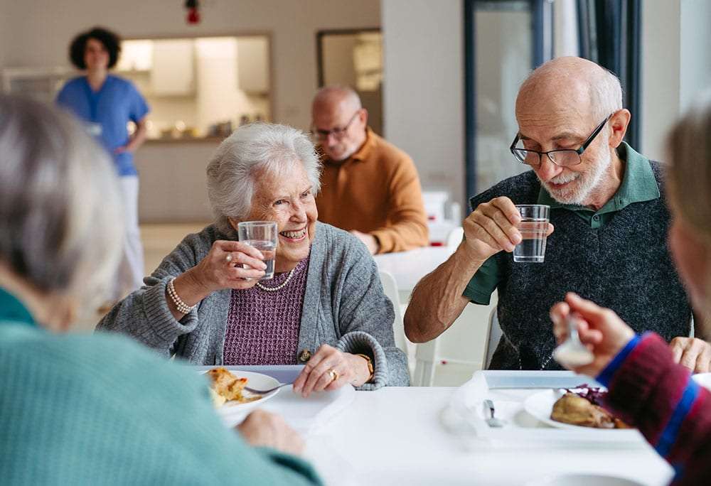 Elderly residents dining together at a senior living facility, with a caregiver visible in the background